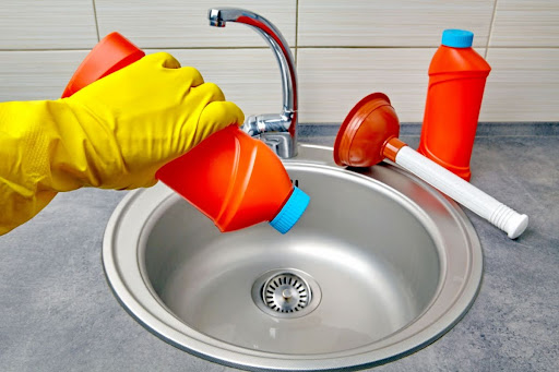 A gloved hand pouring chemical drain cleaner into a sink.