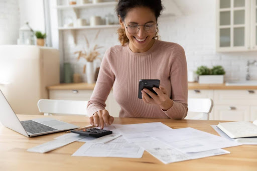 A smiling woman looking at her phone while she pays bills in her kitchen.